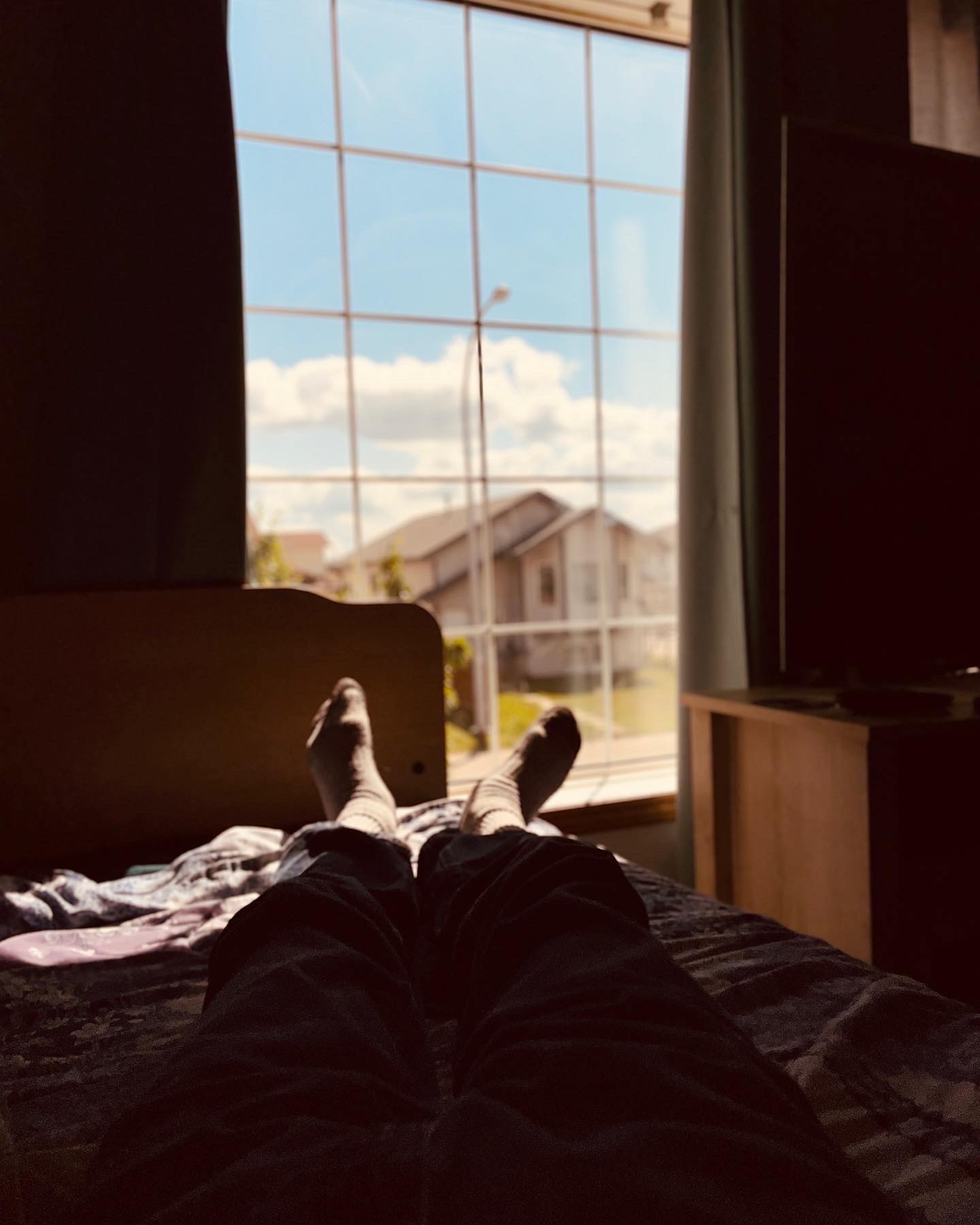 a first-person perspective of an individual reclining on a bed, legs extended in socks and pants, gazing through a sunlit window toward a blue sky with clouds, suburban houses, and green lawn outside. The room appears dimly lit, evoking a sense of quiet contemplation or rest