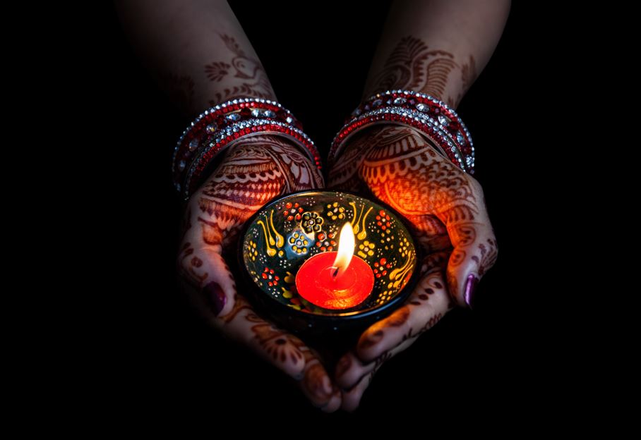 Two hands with henna designs and bangles gently holding a lit traditional diya oil lamp. The warm flame glows on a black background.
